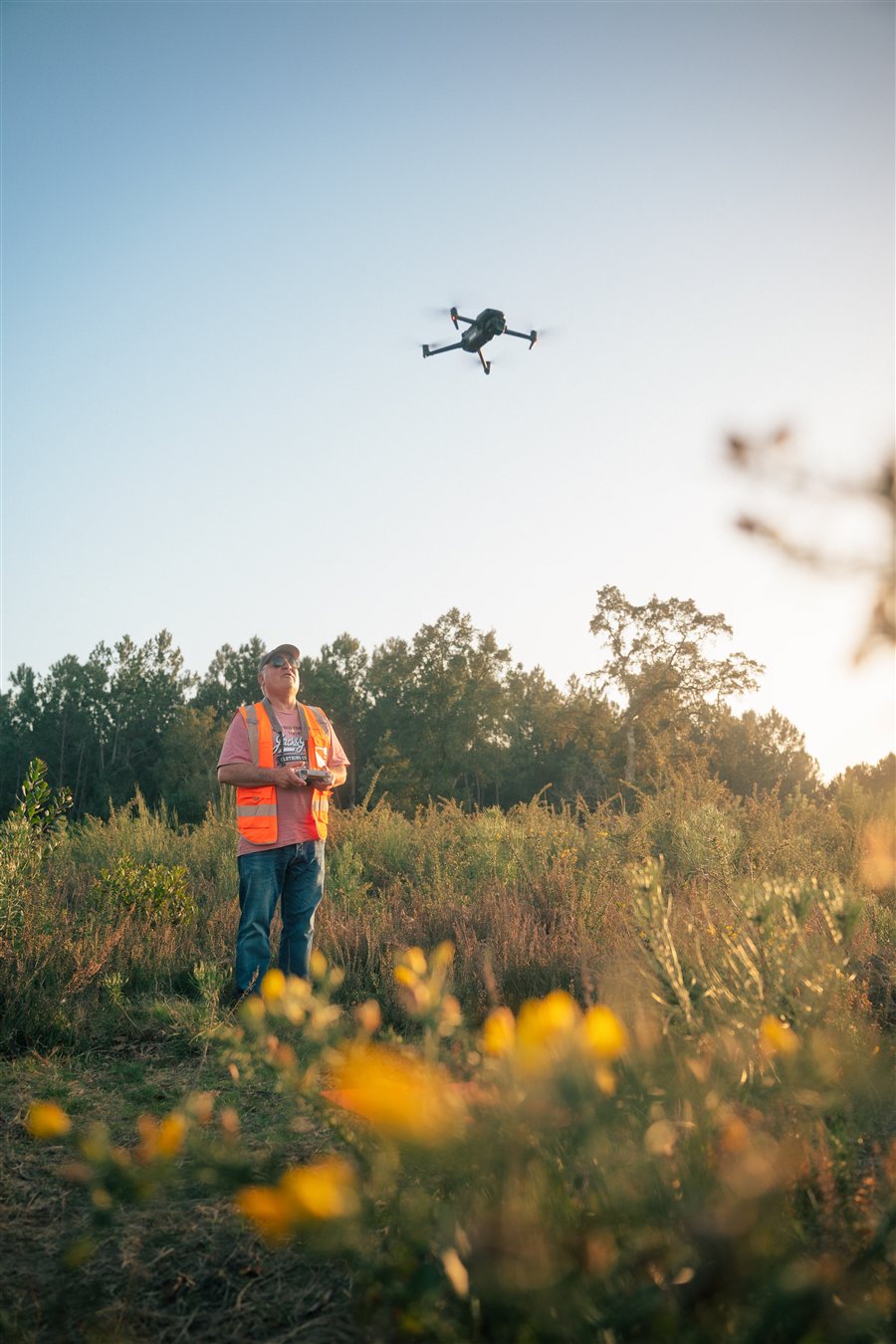 Télépilote en gilet orange, radiocommande en main, qui fait voler un drone au-dessus d'une prairie fleurie.