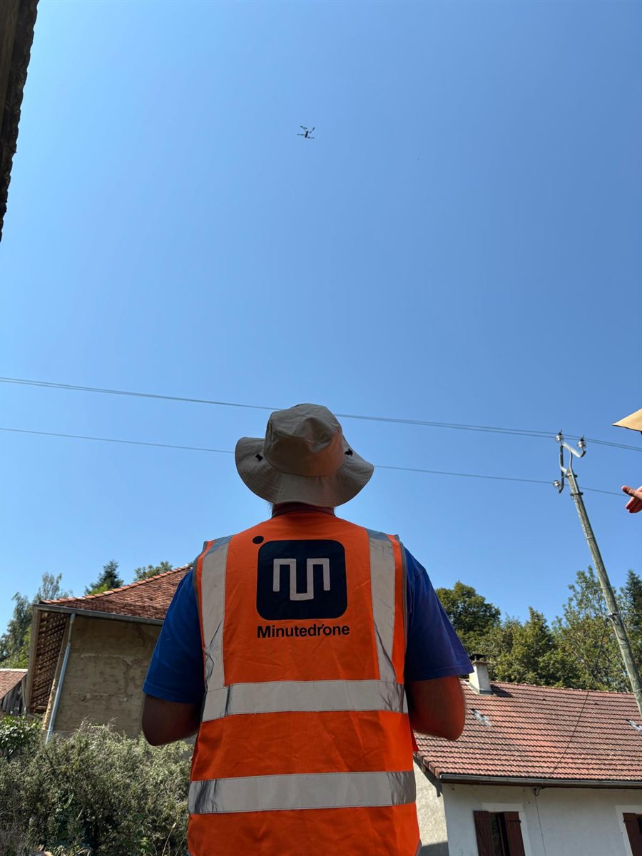 Télépilote de dos, en chapeau et gilet orange Minutedrone, qui suit des yeux un drone en vol dans un ciel bleu, près de maisons.