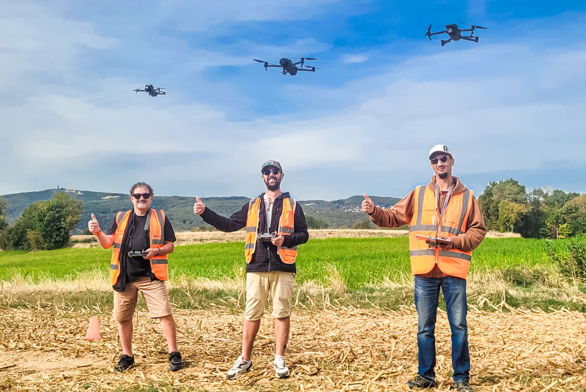 Trois stagiaires en gilet orange, radiocommande en main, pouces levés sous trois drones en vol